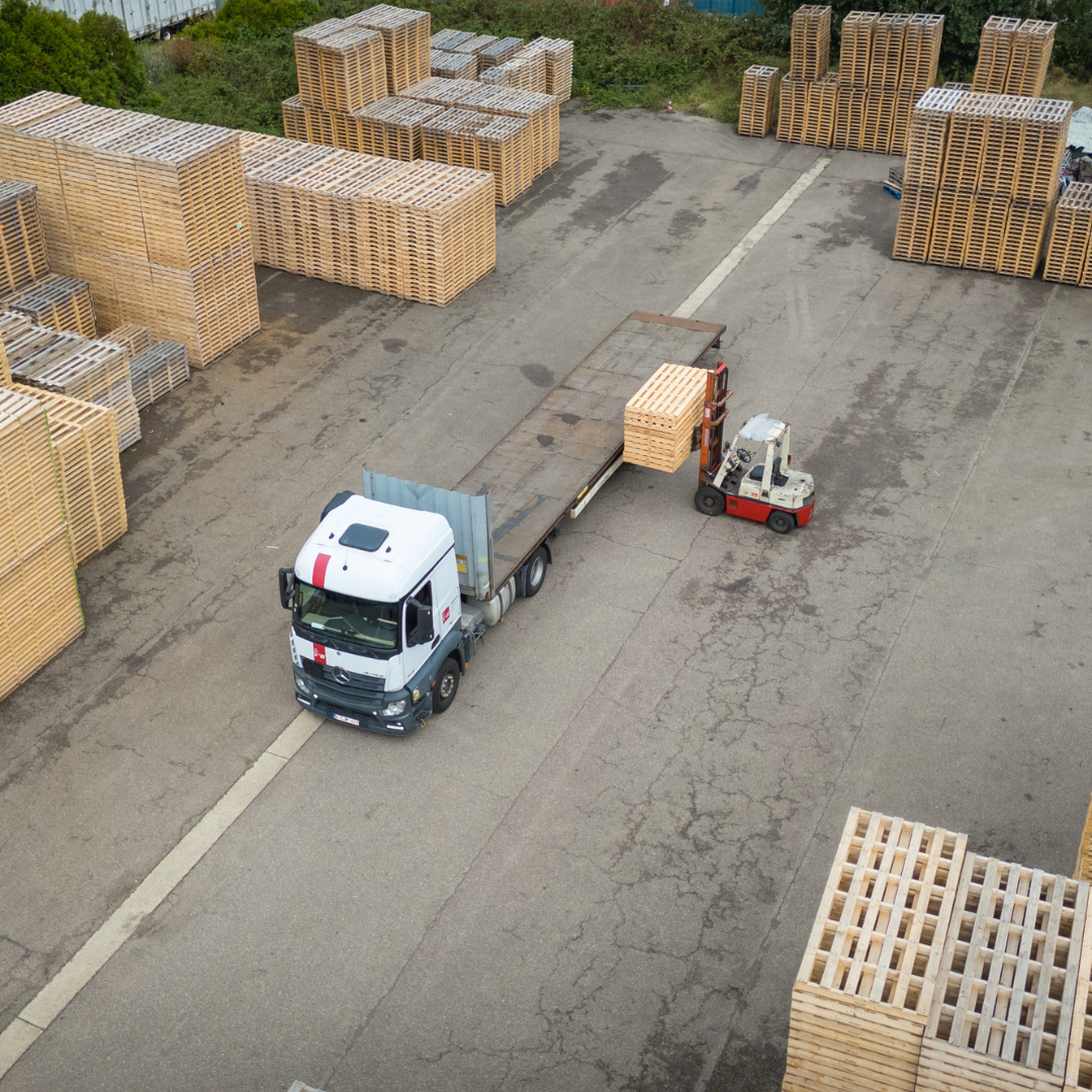 Een heftruck laadt pallets op een vrachtwagen met open laadbak op een houthandel, vanuit de lucht gezien.