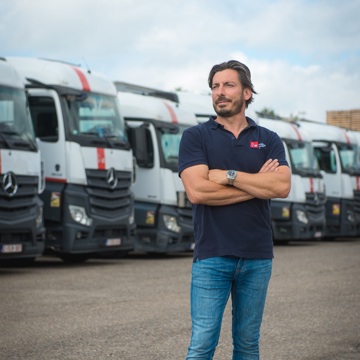 Een man met gekruiste armen staat voor een rij witte vrachtwagens. Hij draagt ​​een donkerblauwe polo en een spijkerbroek.