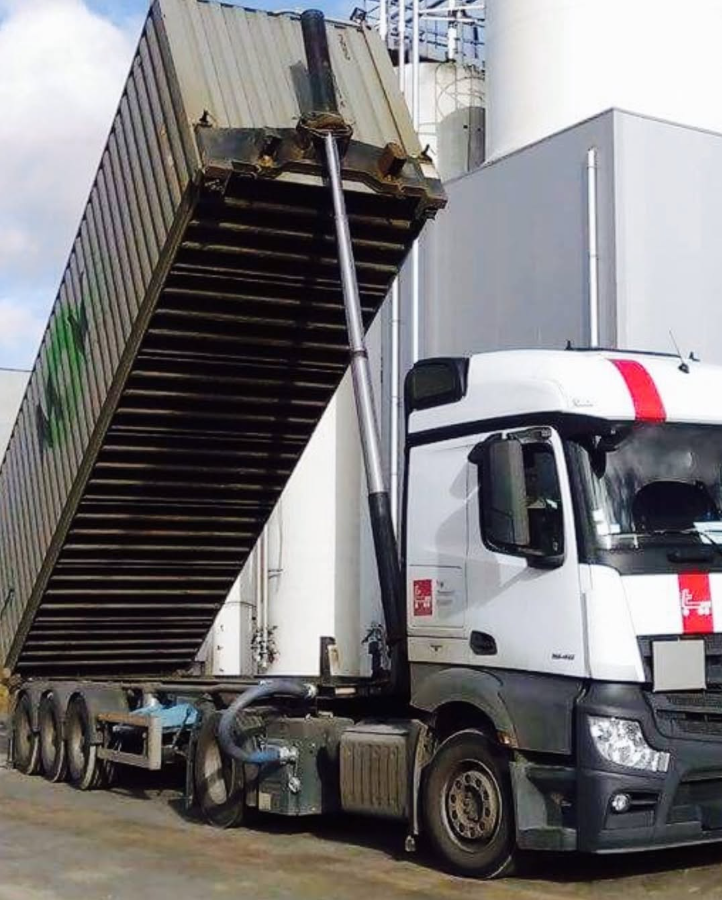 Een vrachtwagen kantelt een zeecontainer, waarschijnlijk om te lossen. Witte vrachtwagen, grijze container, industriële omgeving.