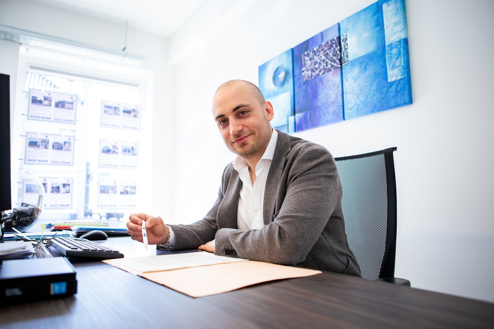 Man aan een bureau, glimlachend, in een kantooromgeving. Grijze jas, wit overhemd, pen in de hand, kijkt naar papieren.