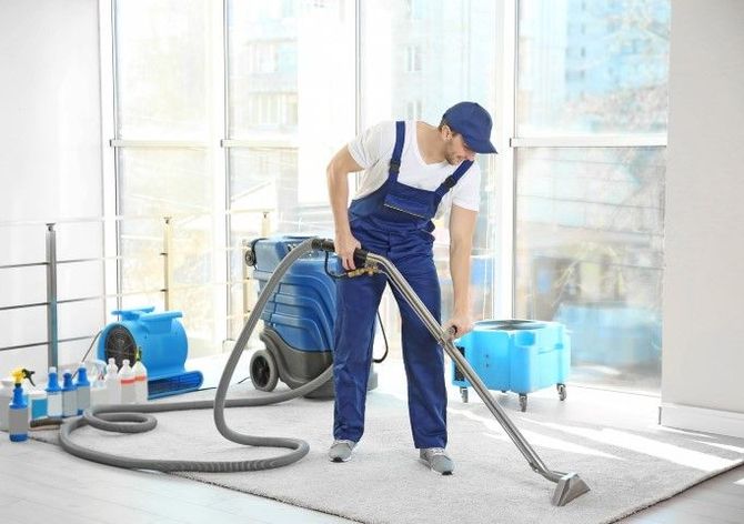 Worker using a floor sander in a bright room, with blue equipment and dust hoses.