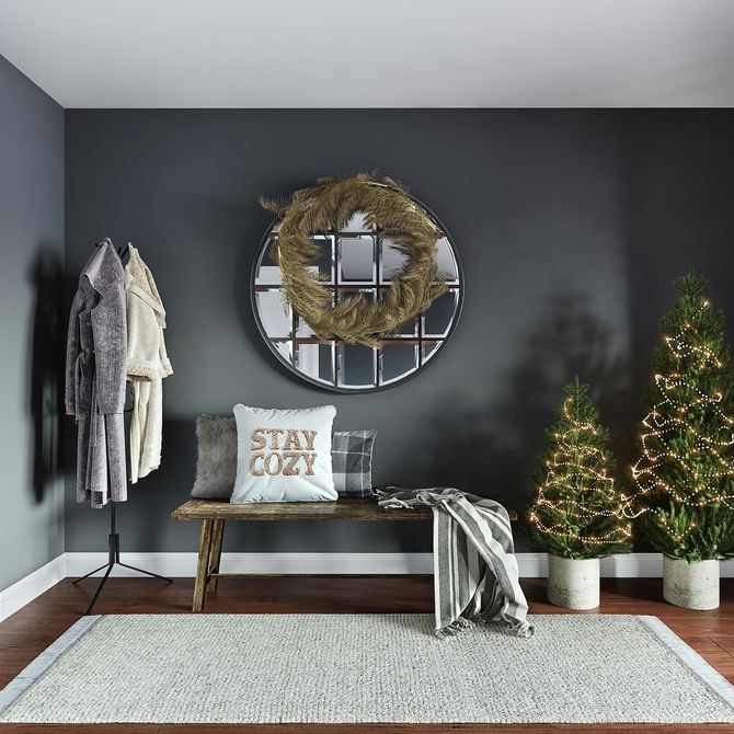 Cozy dark gray entryway with bench, wreath mirror, coat rack, and lit Christmas trees on a white rug