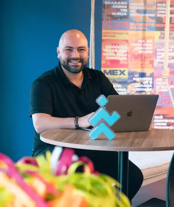 Man with a beard smiles, sitting at a table with a laptop and a blue logo. Colorful wall background.