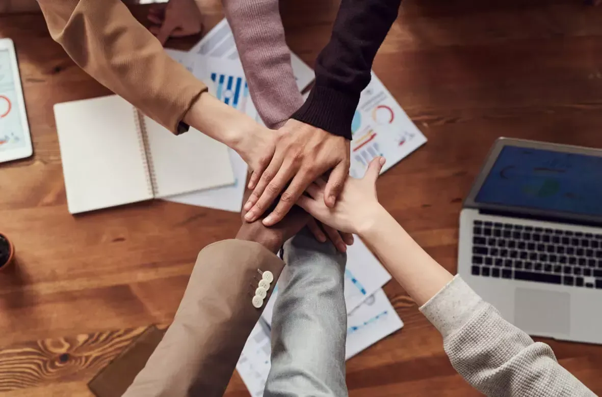 Hands stacked together in a team huddle over a wooden table, with charts and a laptop visible.