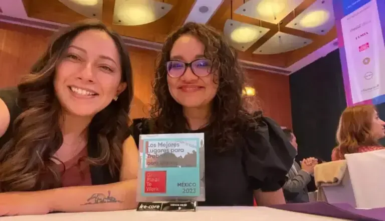 Two smiling women seated at a table, holding a small sign at a conference or event.