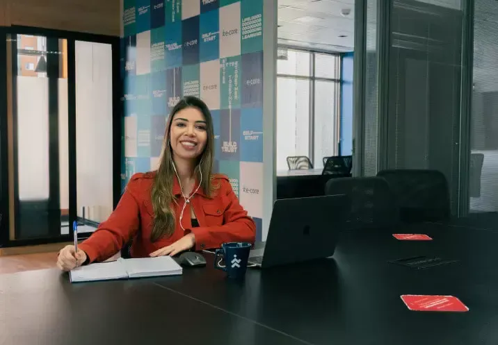 Woman in red jacket smiles at a table with a laptop, notebook, and a mug in an office setting.