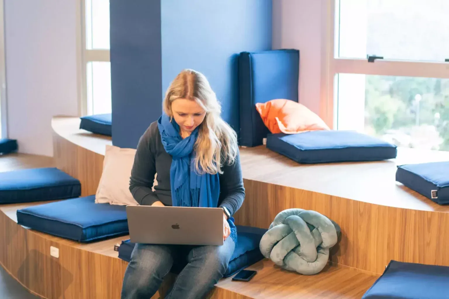 Woman sitting on wooden seating, working on a laptop. Blue scarf, pillows, and modern office setting.