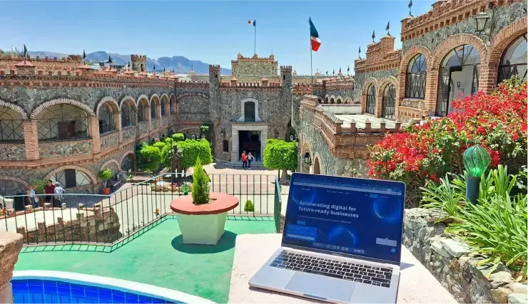 Laptop on a patio overlooking a stone castle with blue pool and green plants.