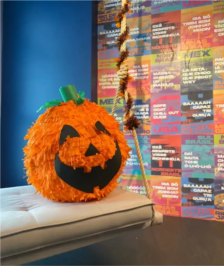 Orange pumpkin piñata with a smiling face, resting on a white bench. Brown and gold garland hangs nearby.