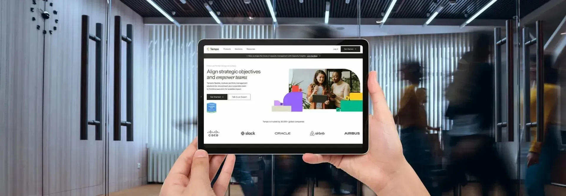Hands holding a tablet displaying a website. People are blurred in the background, walking near glass doors.