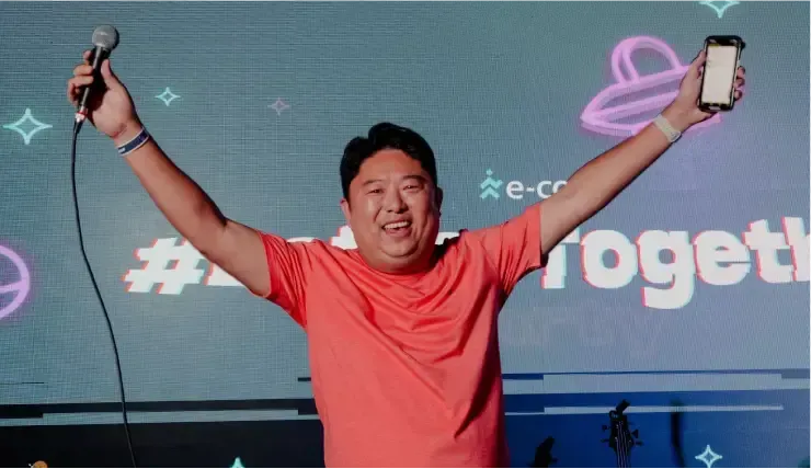 Man in red shirt holds a phone and microphone, arms raised in a celebration on a stage with a screen.