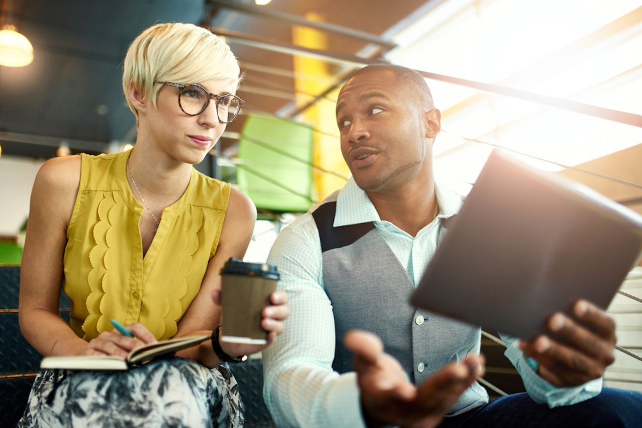 Two people discussing on stairs, one holds a tablet, the other a notebook and coffee.