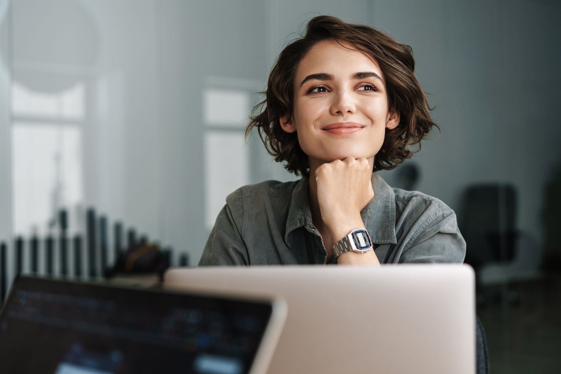 Woman smiling, resting chin on hand, looking upwards, near laptop and tablet.