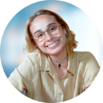 Woman with glasses smiles; wearing a beige shirt and a dark pendant necklace against a blue background.