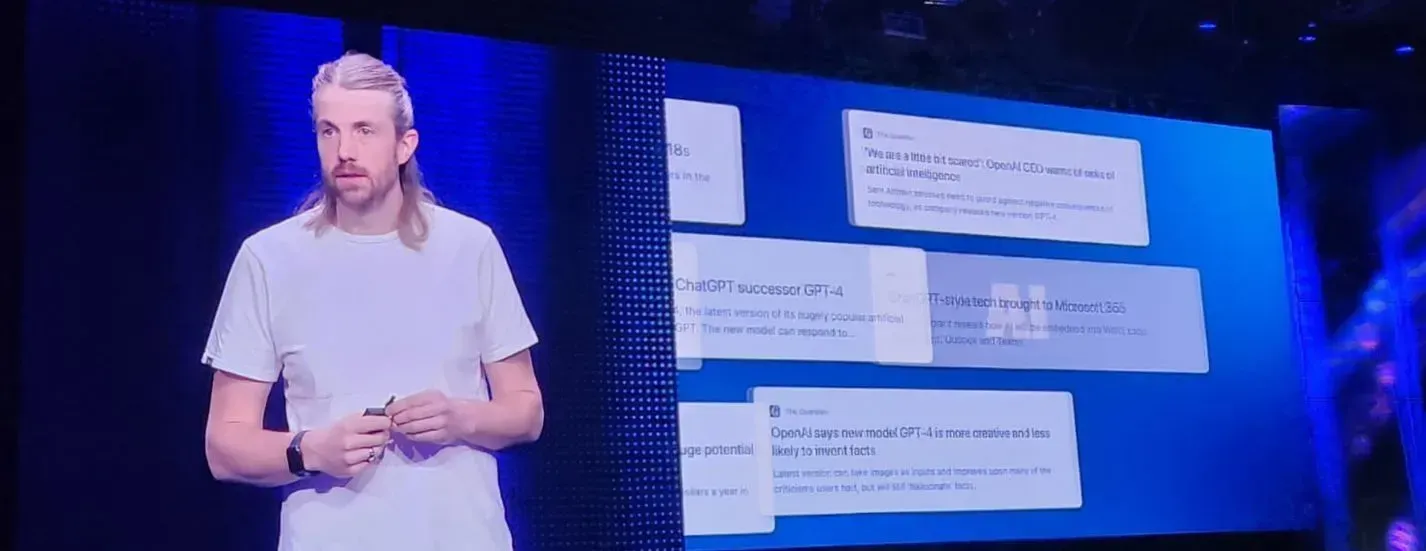 Man with blonde mullet speaks at a presentation, screen with text in the background.