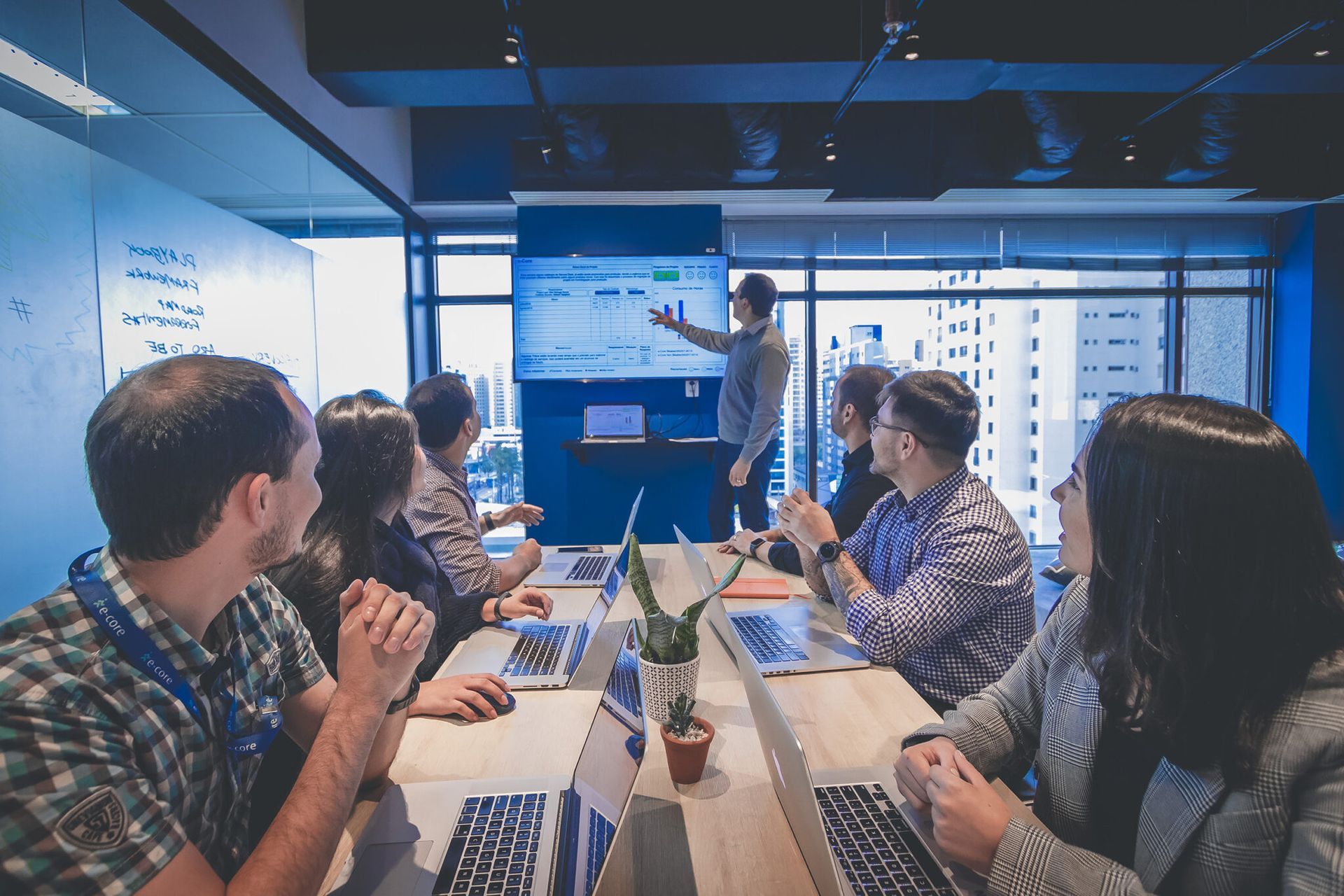 People in a meeting room, a man points at a screen with charts. Others clap. Blue and modern office.