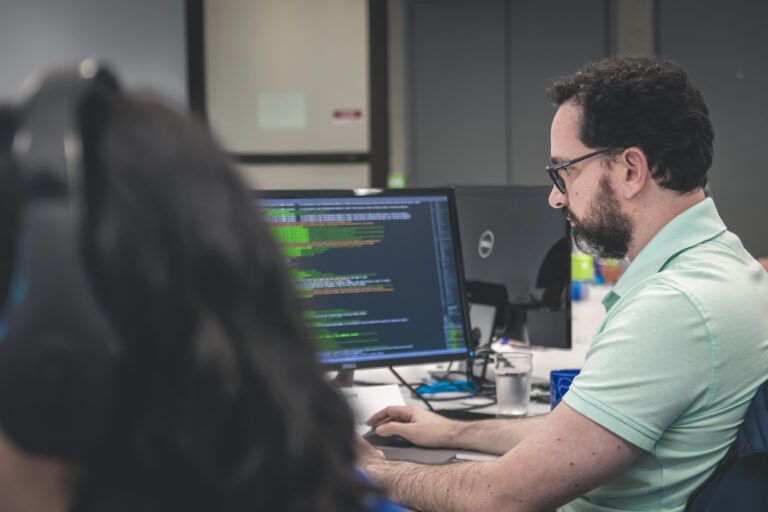 Man with glasses working on a computer at a desk, looking at code displayed on the screen.
