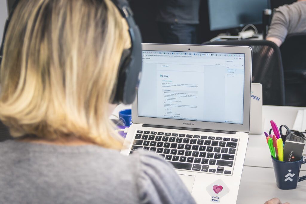 Woman with blonde hair wearing headphones works on a laptop at a desk. Pens in a cup are visible.