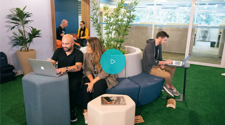 People working on laptops in a modern office lounge area. Some seated on blue ottomans, others in the background. Green decor.