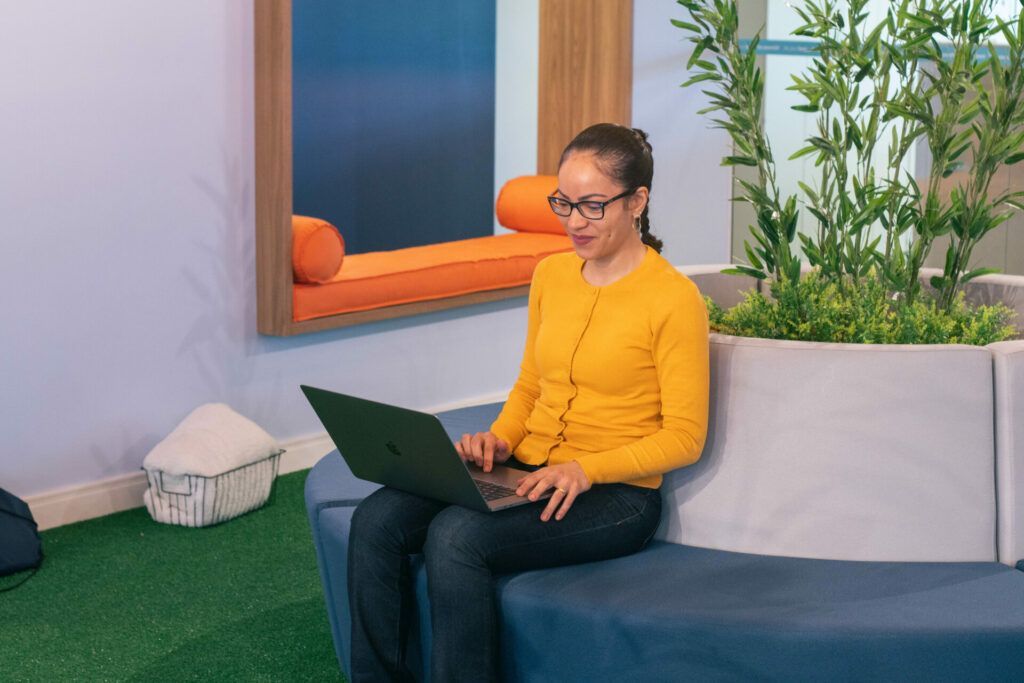 Woman in yellow top, glasses, using laptop, seated on blue couch, near green plants.