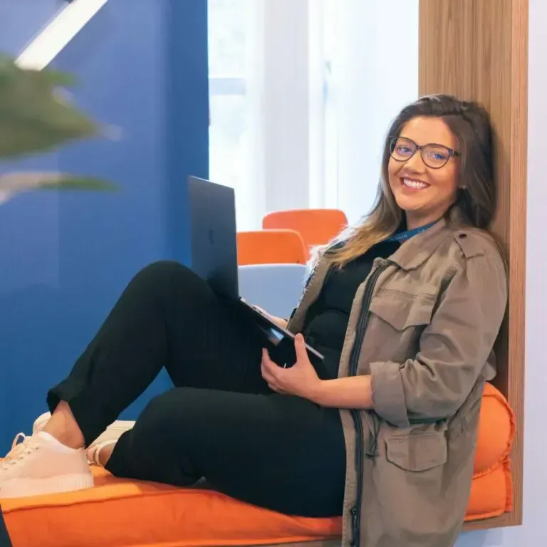 Woman in glasses sits with a laptop on orange bench, smiling, wearing a jacket in a modern office.