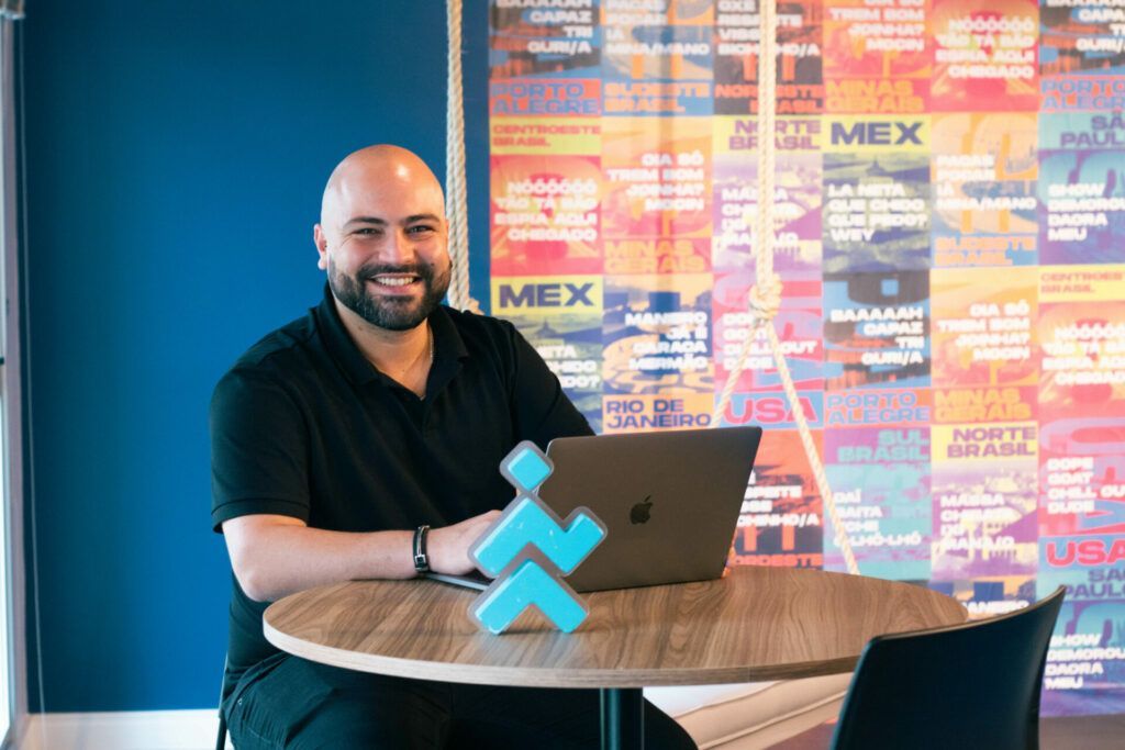 Man smiling, sitting at a table with a laptop, logo, and a colorful mural in the background.