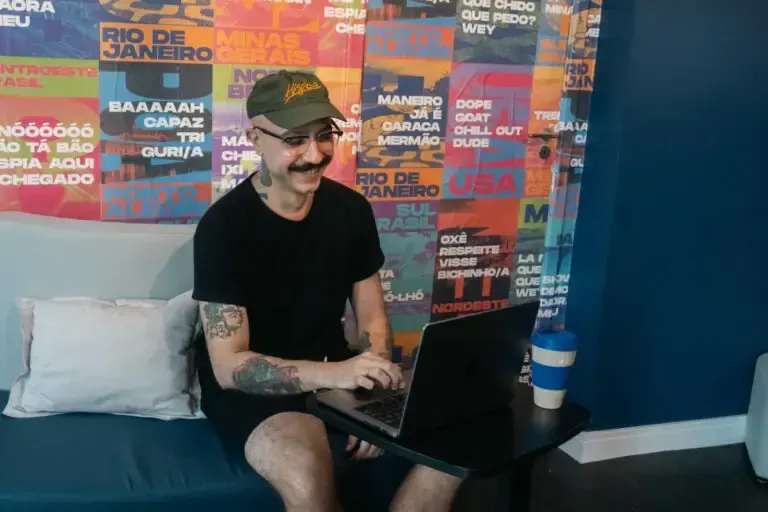 Person with tattoos and mustache using a laptop, sitting on a blue couch in front of a colorful wall with text.