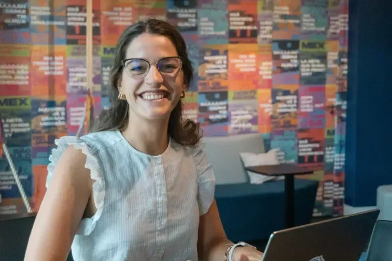 Woman with glasses smiles at the camera while working on a laptop in a colorful office setting.