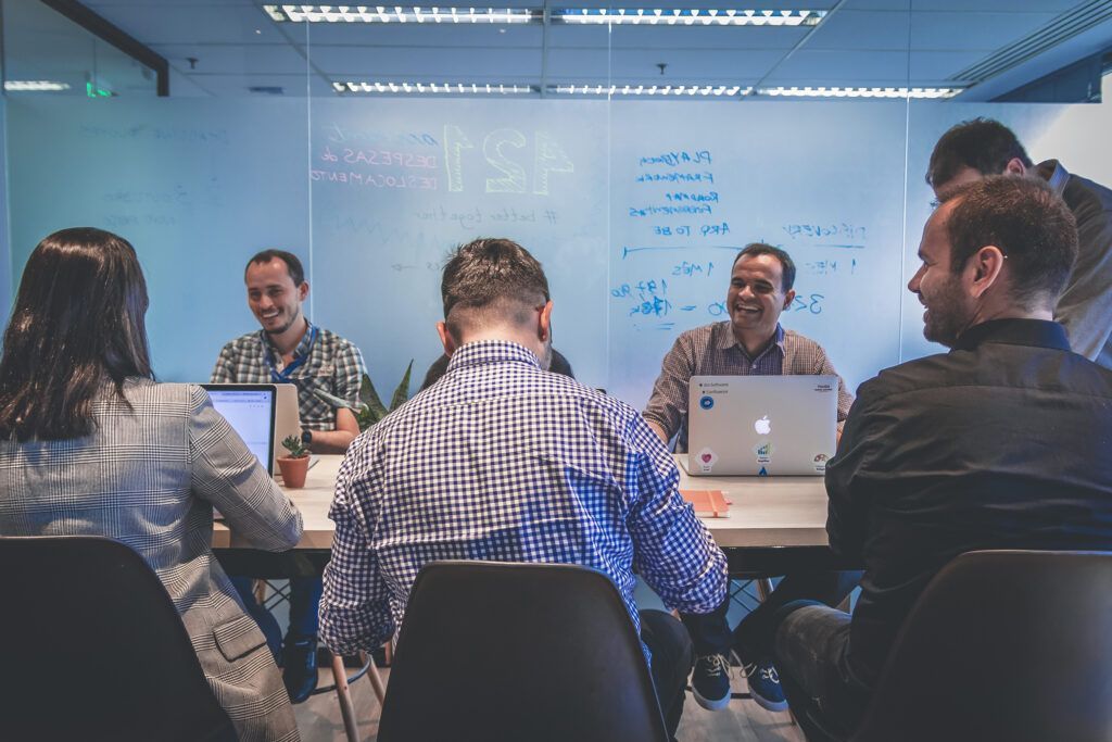 Group of people collaborating around a table, whiteboard in background, laptops visible, some smiling.