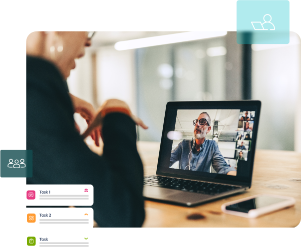 Woman in a blazer on a video call, looking at a laptop with multiple faces. Phone and task list on the desk.
