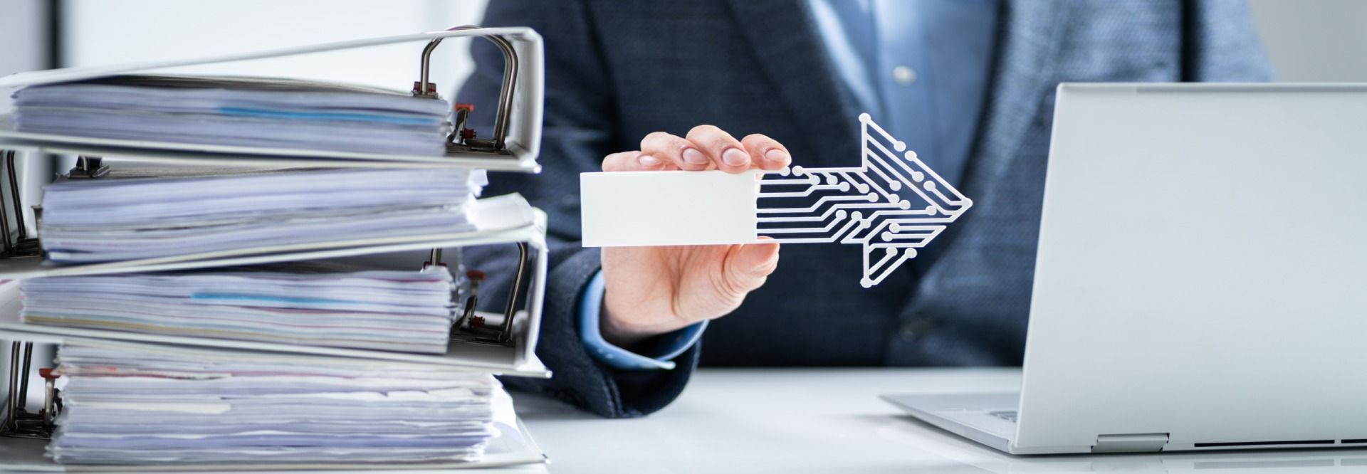 A person holding an arrow-shaped circuit board, next to a stack of papers and laptop.