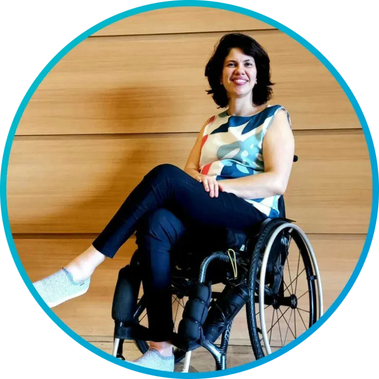 Woman in wheelchair smiles, wearing a patterned top and black pants, in front of a wooden wall.