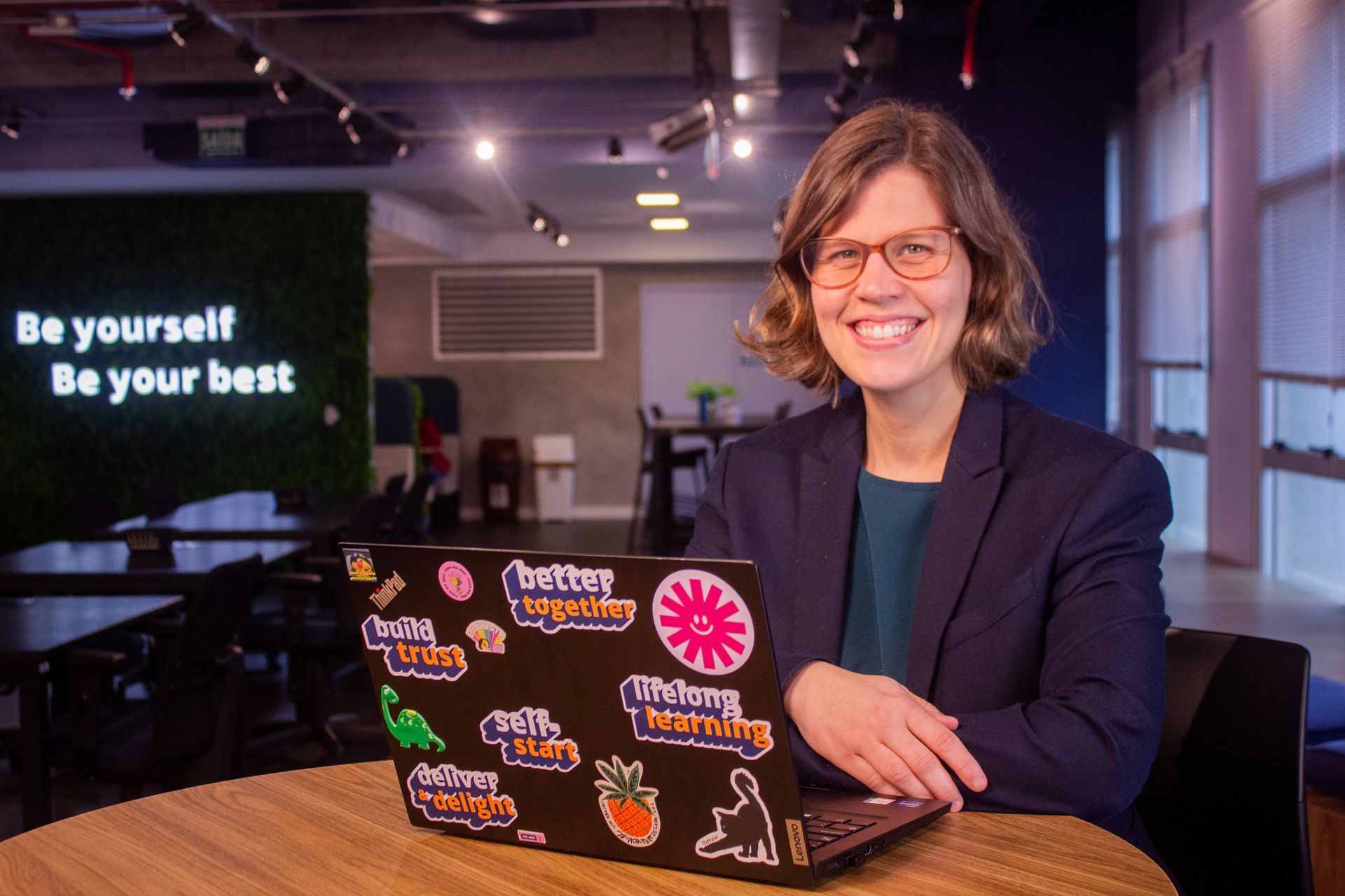 Woman in red jacket smiles at a table with a laptop, notebook, and a mug in an office setting.