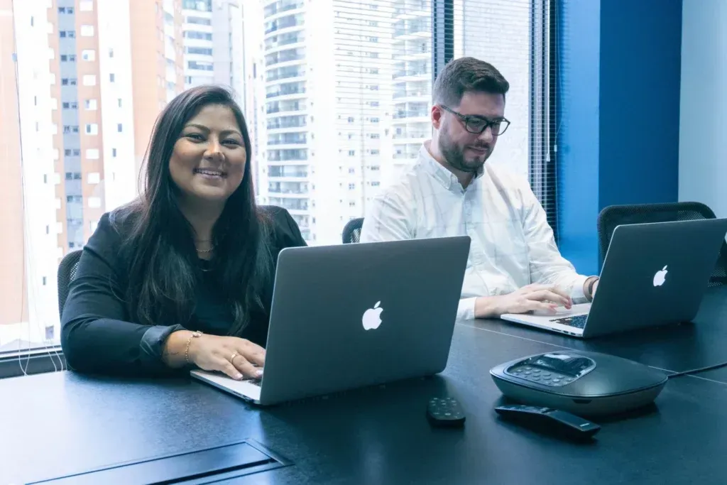 Two people working on laptops at a table in an office, smiling.
