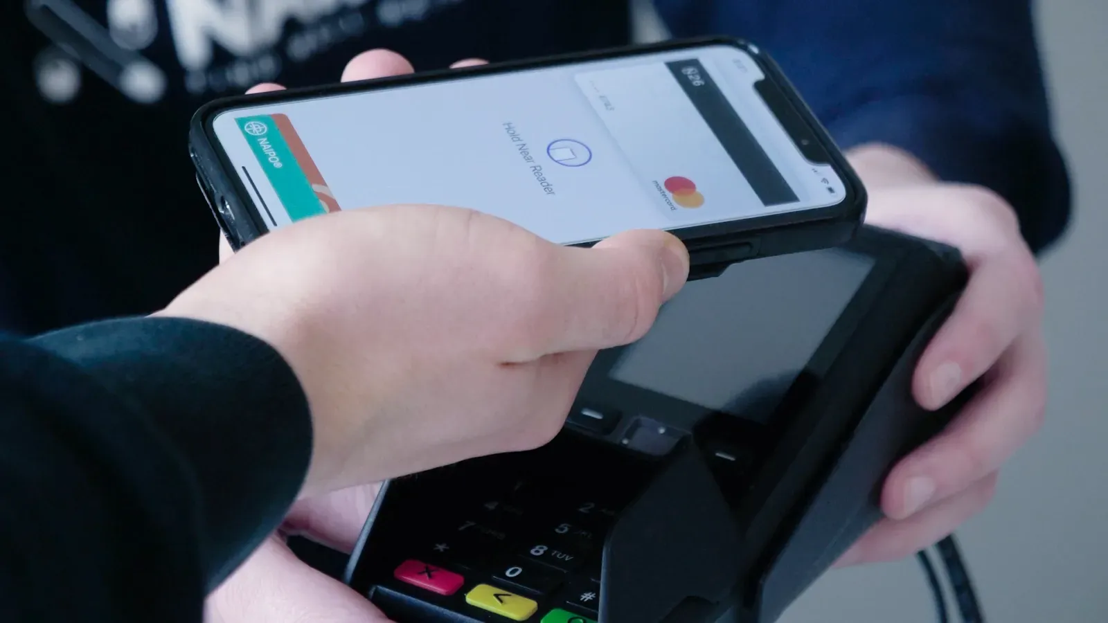 Person paying with phone held over a payment terminal. Screen shows payment processing, credit card image.