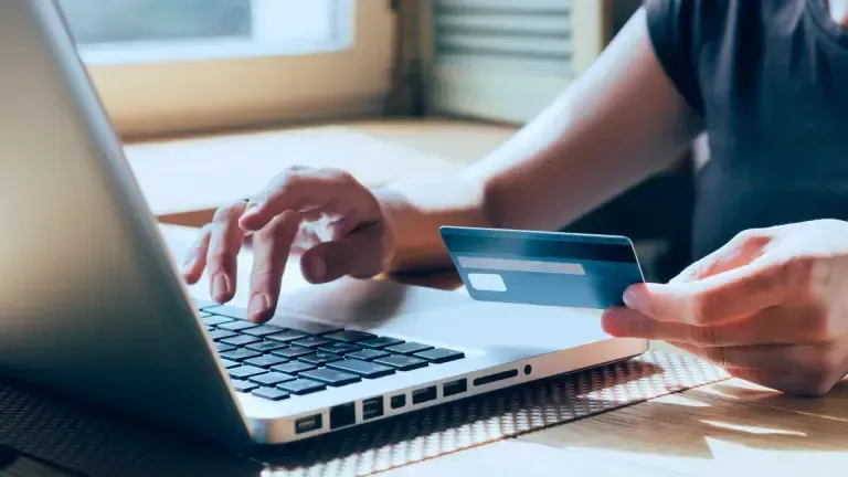 Person using a laptop, holding a credit card. Fingers typing on keyboard indoors near a window.
