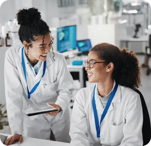 Two people in lab coats laugh, looking at a tablet in a lab setting.