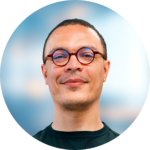 Man with glasses smiles, set against a blurred blue background.