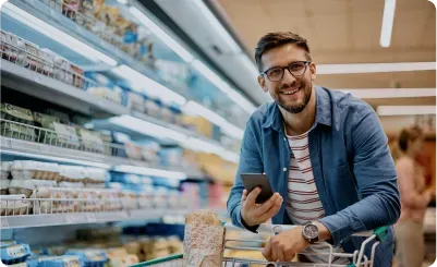 Man in glasses smiling, holding a phone, leaning on a shopping cart in a grocery store.