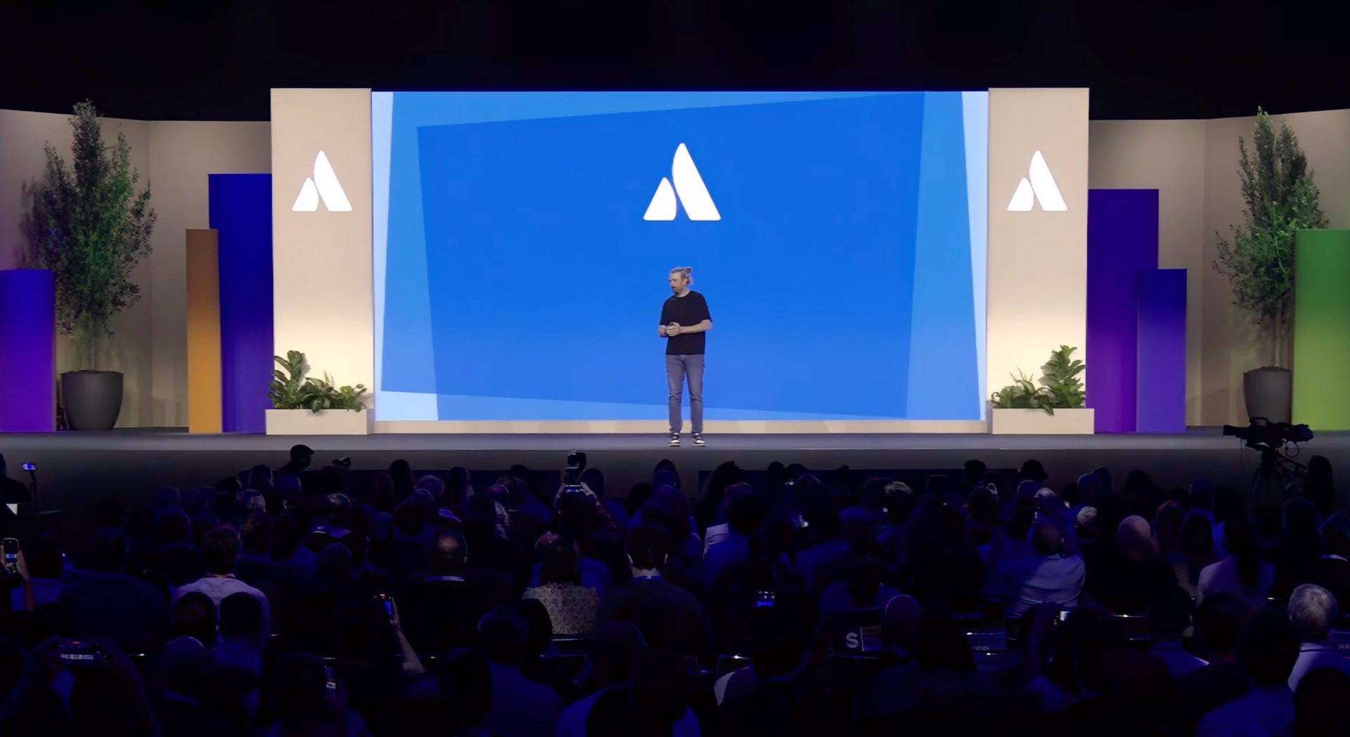 Man on stage with a logo behind him, presenting to a large audience.