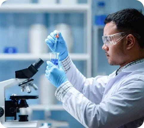 Scientist in a lab coat and gloves adding liquid to a beaker, wearing safety glasses. Microscope visible.