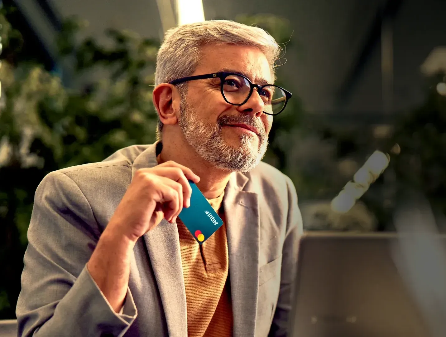 Man in glasses, gray hair, holding a teal credit card, smiling, sitting near a laptop.