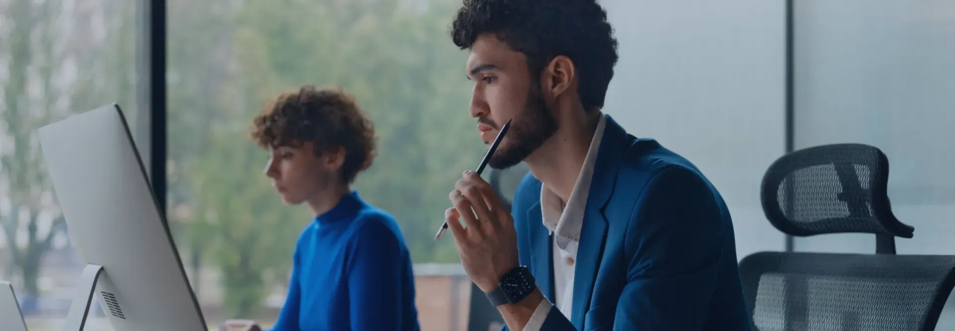 Two people at desks near a window; one man holding a pen to his mouth, deep in thought.