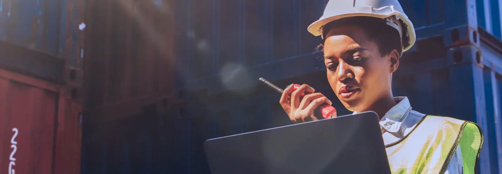 Woman in hard hat and vest, using a laptop and walkie-talkie near shipping containers.