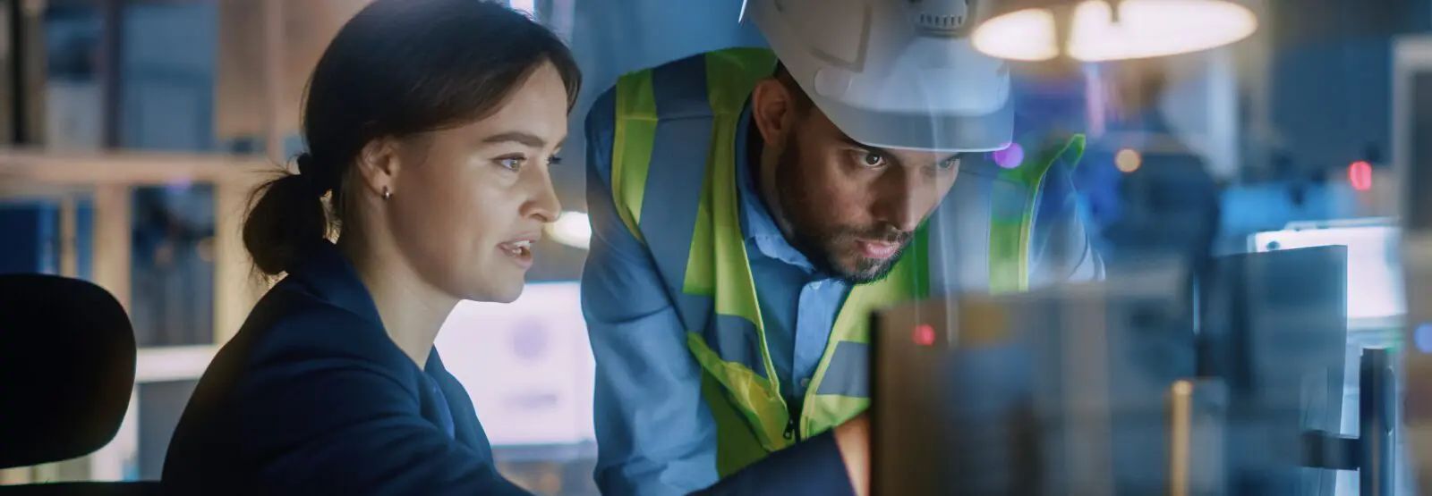 Two people looking at a computer screen, one wearing a safety vest and hard hat.