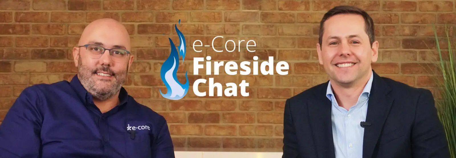Two men in suits seated in front of a brick wall, logo