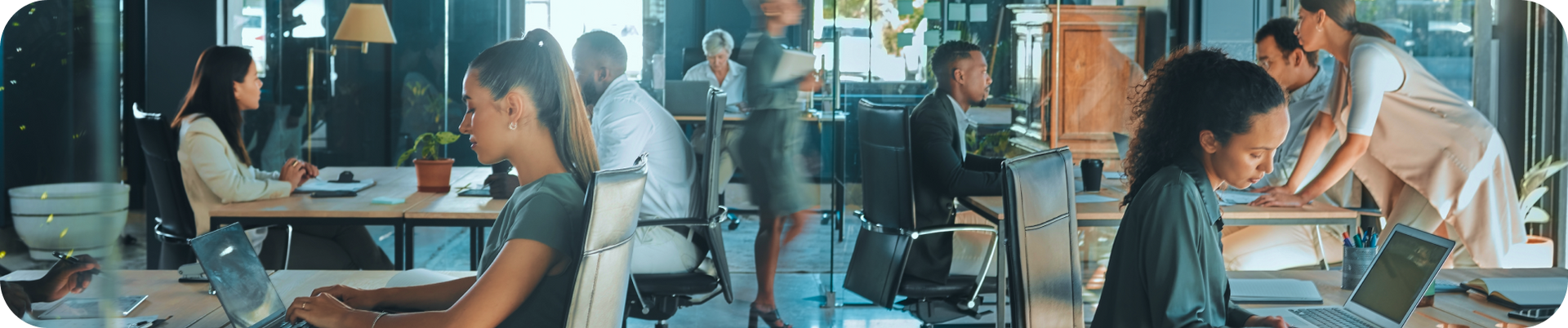 People working at desks in a bright office. One woman types on a laptop. Others stand or sit.