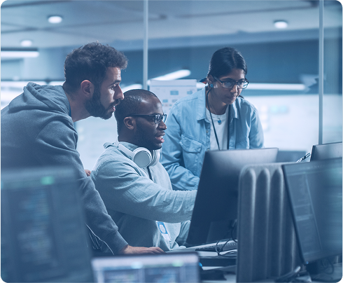 Three people collaborating at computer monitors in a tech office, focused intently.