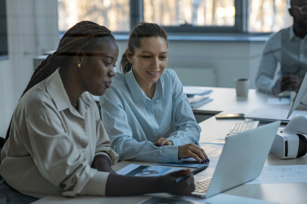 Two women looking at a laptop, one points at the screen. Office setting, daylight.