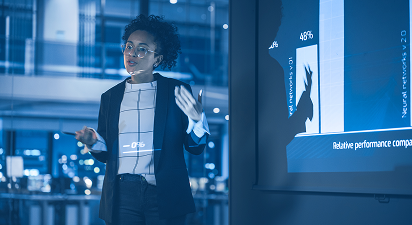 Businesswoman presenting data on a screen in a modern office setting, gesturing with hands.